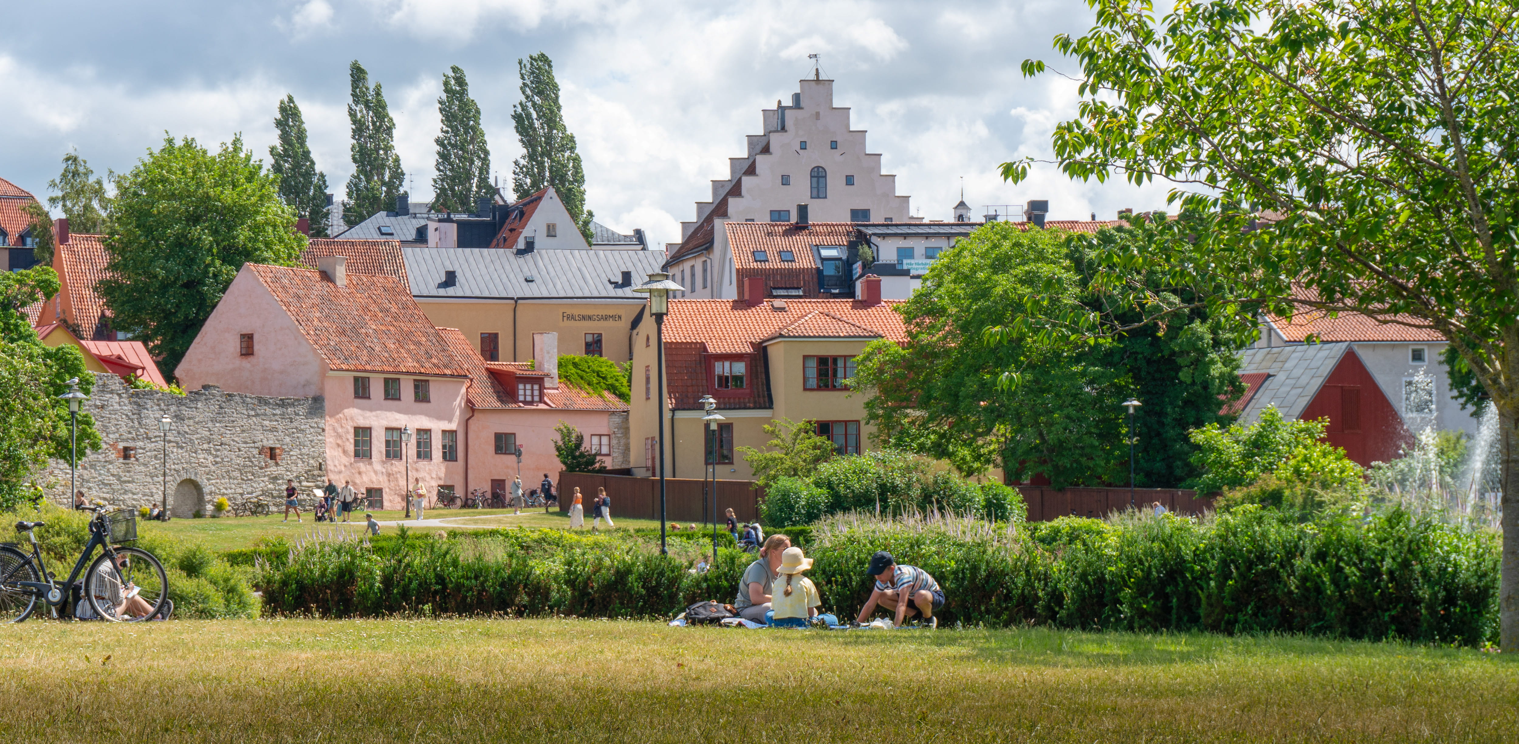 En solig sommardag i Almedalen i Visby, med människor som picknickar i parken framför färgglada gamla hus och grönskande träd.